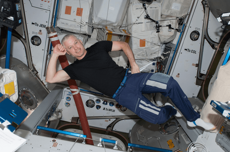 Swanson is a white man. He's floating in a reclined position wearing a black t-shirt, blue sweatpants, and white socks