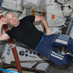 Swanson is a white man. He's floating in a reclined position wearing a black t-shirt, blue sweatpants, and white socks