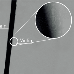 A photo showing a microscope image of a hair with the tiny violin next to it. In the main image, the violin is not actually visible next to the comparatively enormous hair that cuts through the centre of the image. The hair is effectively a black line in the middle of the picture, but there is a zoomed in bubble to the top right of the image showing the violin close up.
